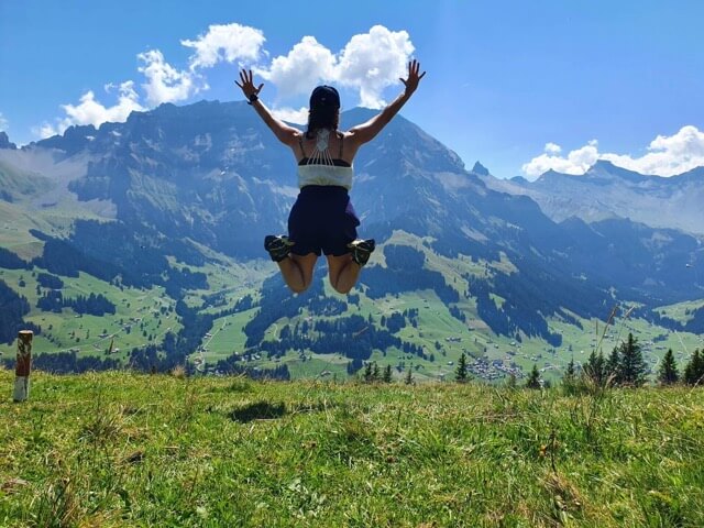 portrait d'une femme de dos en train de sauter dans les airs face à un paysage montagneux en plein soleil.